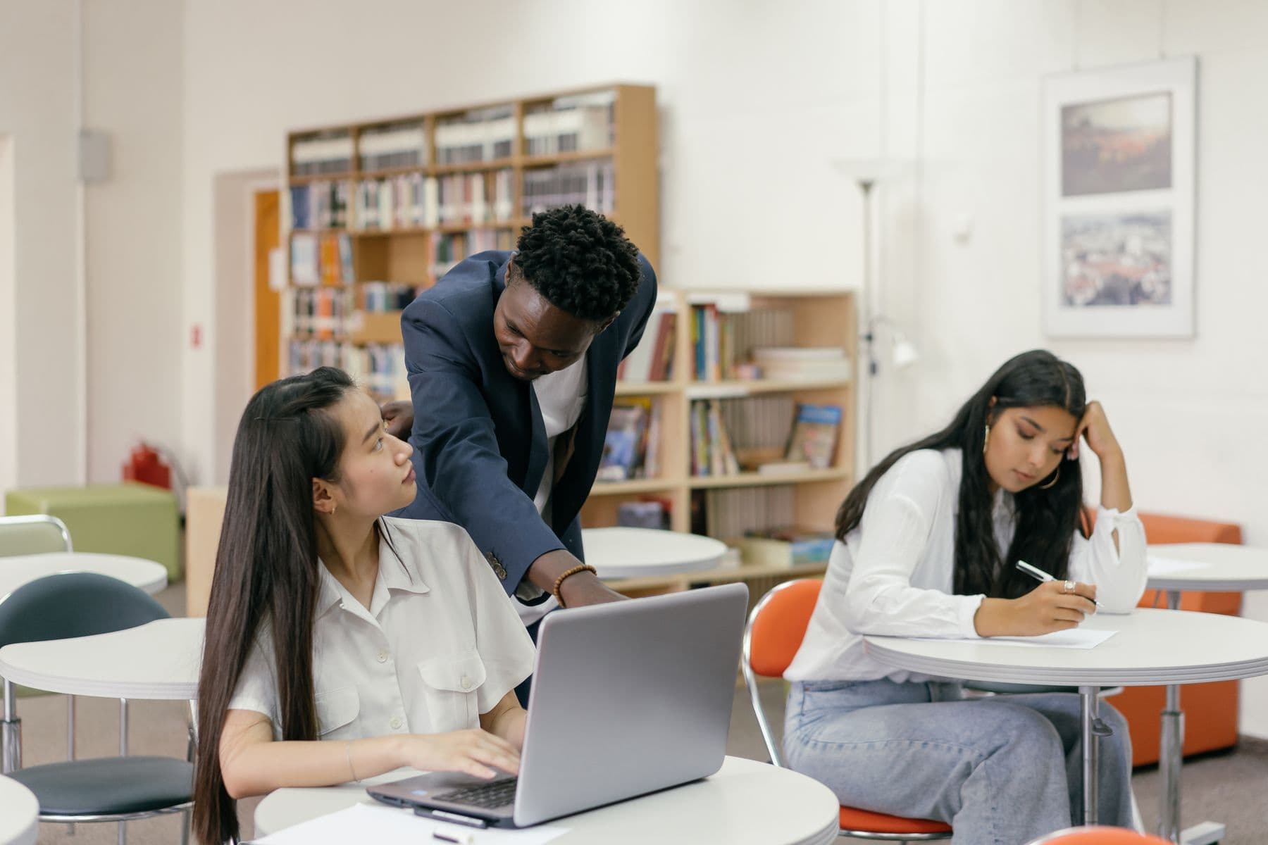 An educator leans over to point at a student laptop in a bright library, with another student writing nearby.