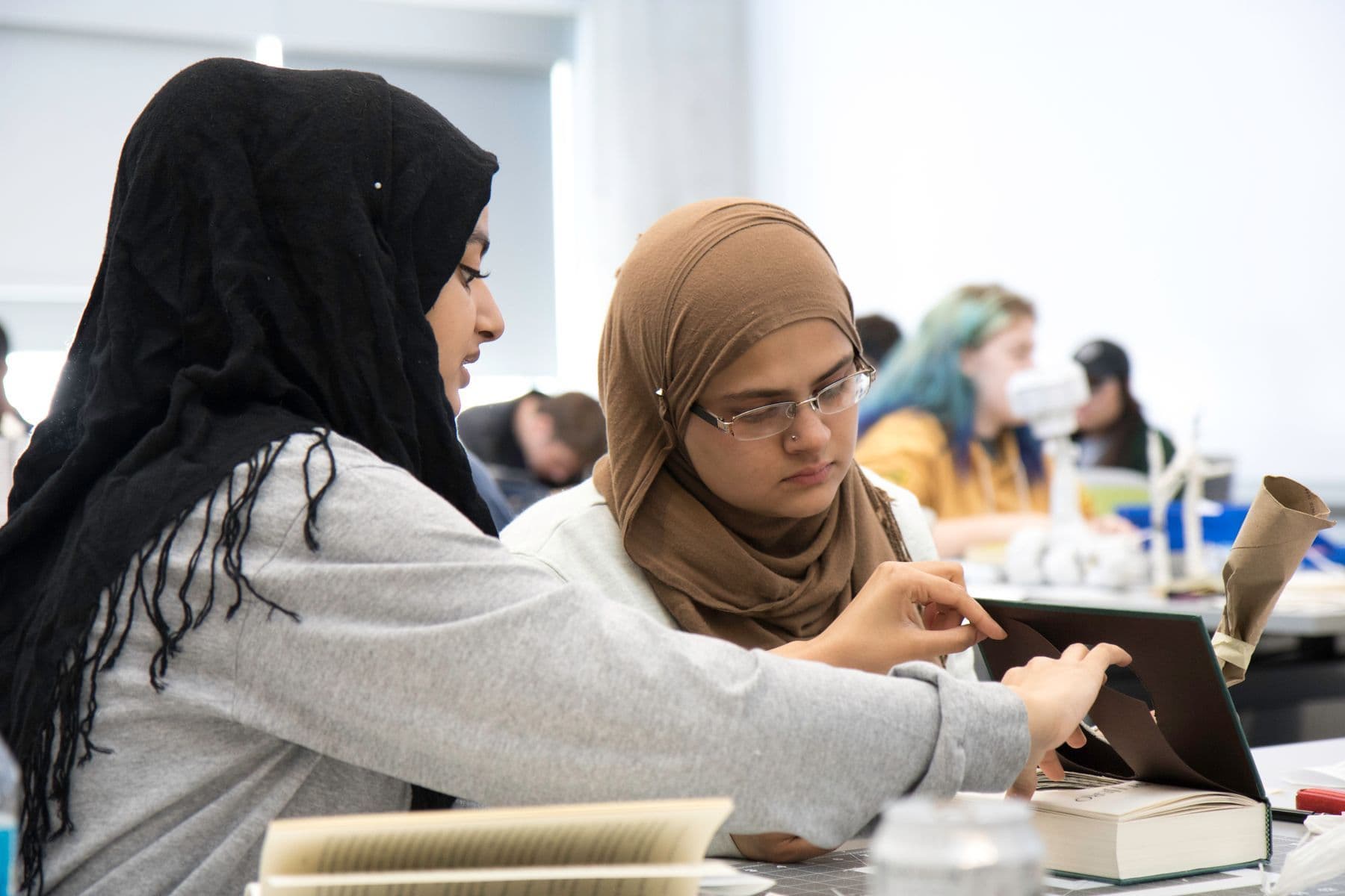 Two women in headscarves sit side by side at a table, one pointing at a laptop while they work together.