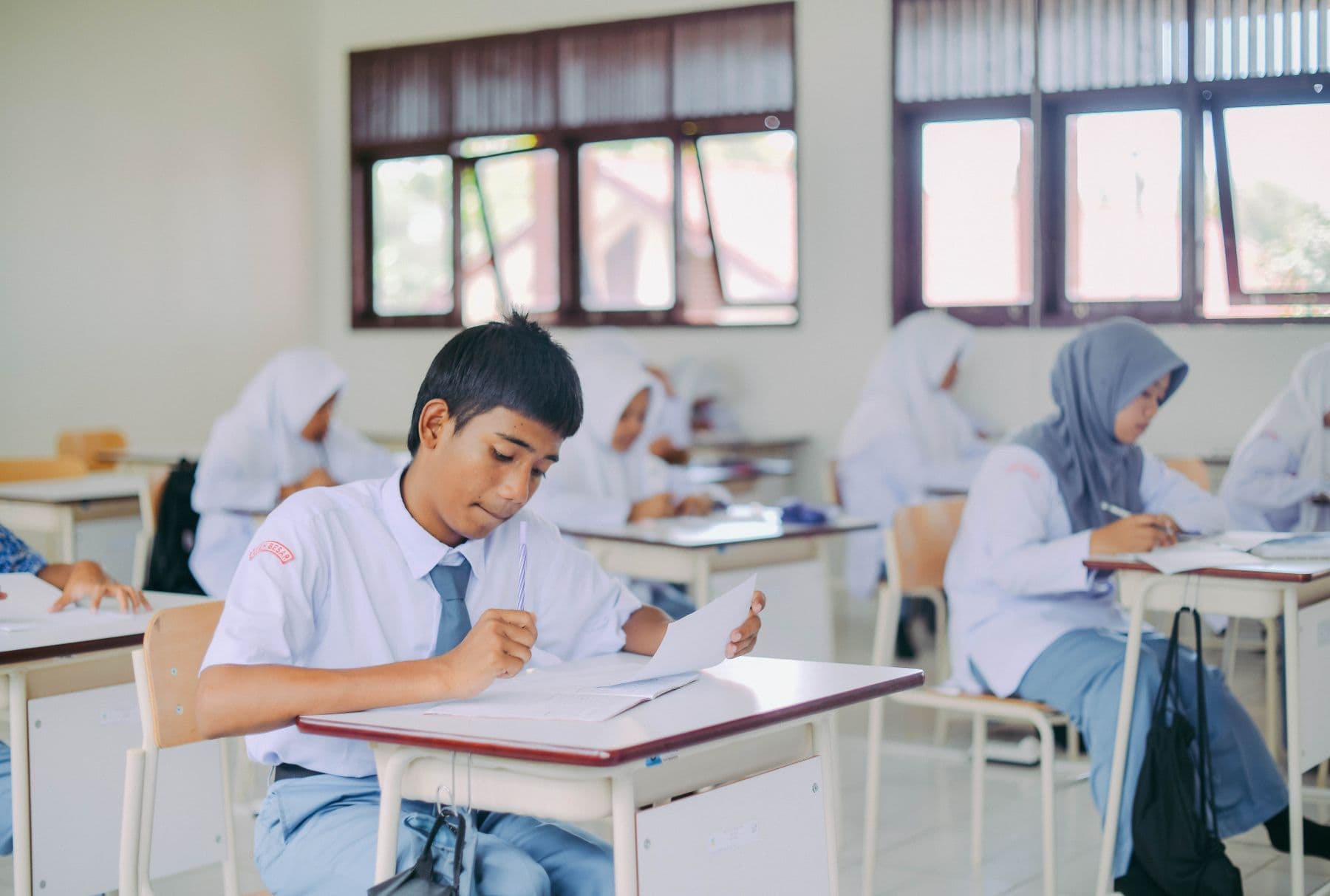 High school students in white uniforms write at their desks in a bright classroom with tall windows.