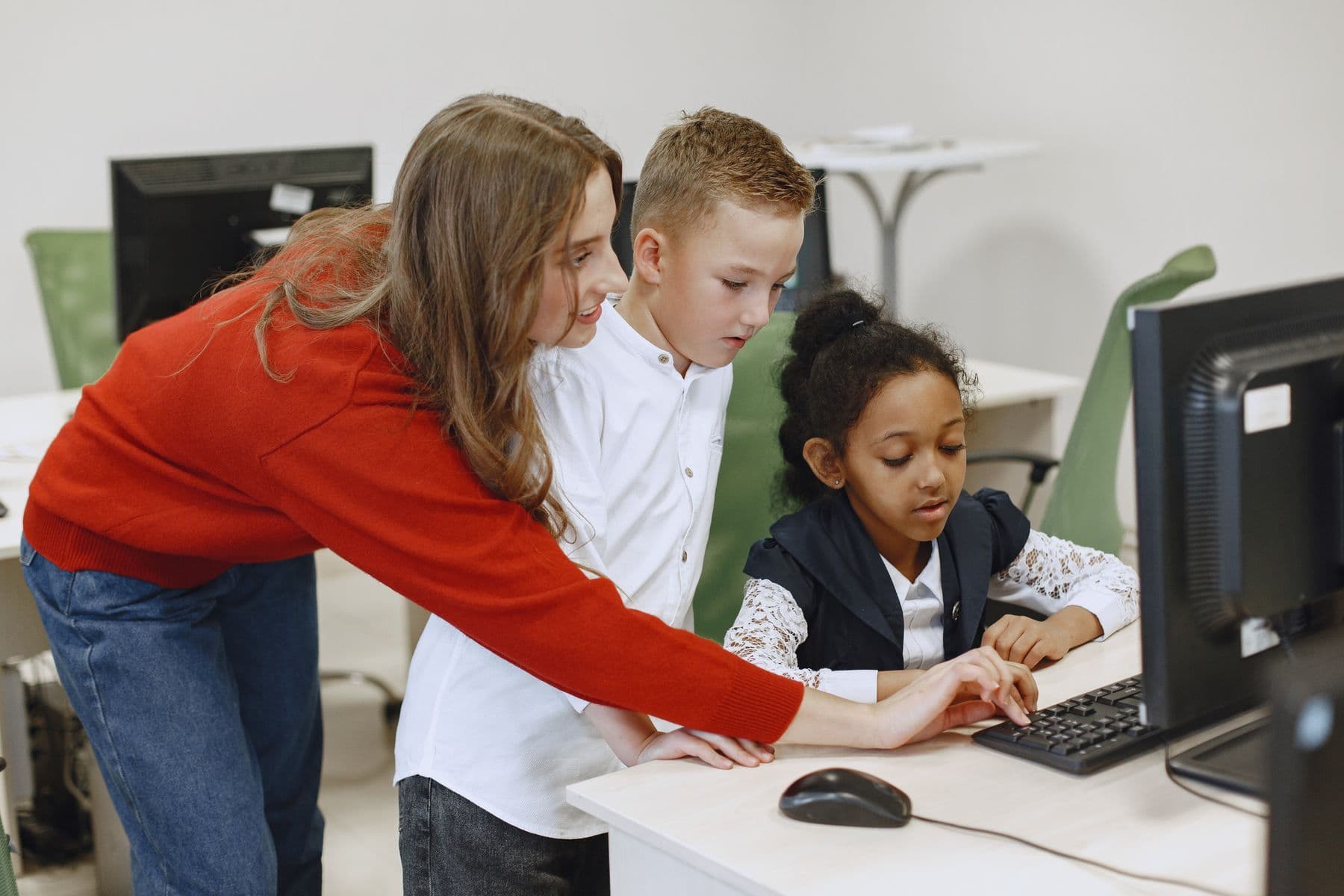 An educator in a red sweater leans in to help two young learners at a desktop computer.